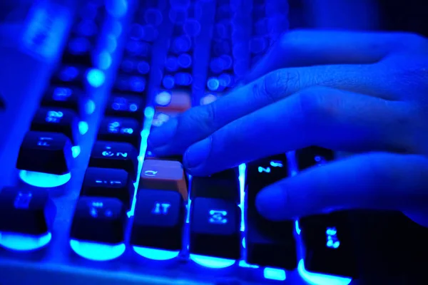 A hand pressing keys on a backlit mechanical keyboard glowing bright blue in the dark
